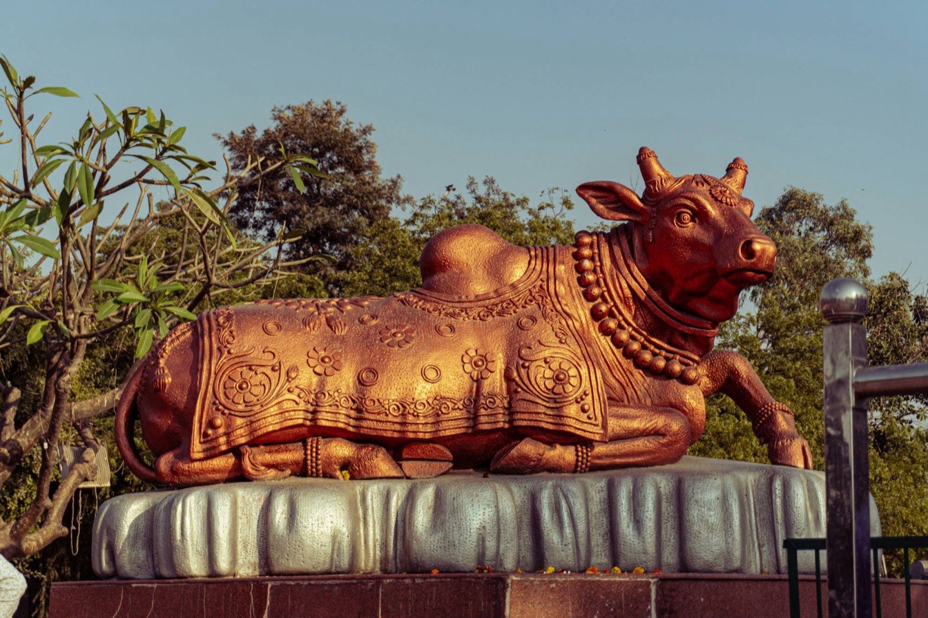 A painted stone statue of Nandi, the bull, reclining on a carved plinth and garlanded with strings of bells, under an open sky with trees behind.