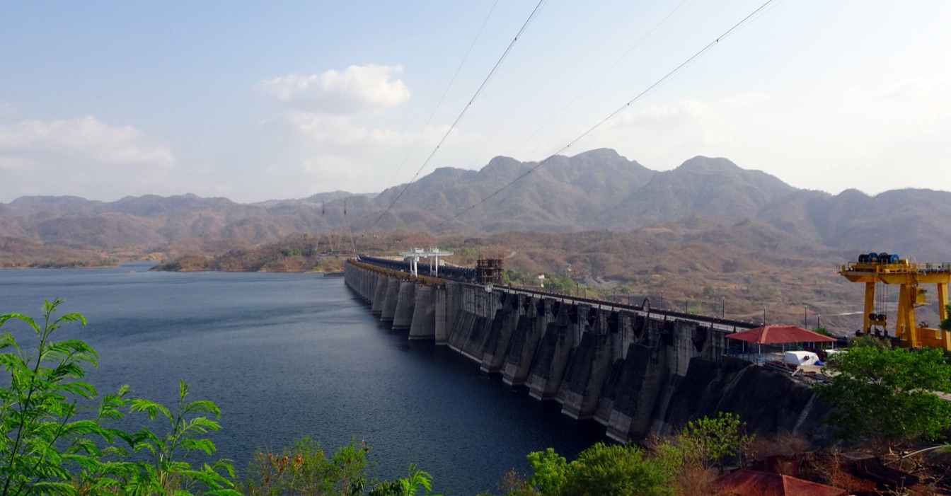 A long concrete gravity dam with a row of buttressed spillway piers runs from the foreground into the distance, holding back a wide dark reservoir on the left. Dry, scrub-covered hills rise behind it; a yellow gantry crane stands at the right end of the crest and power lines cross the sky.