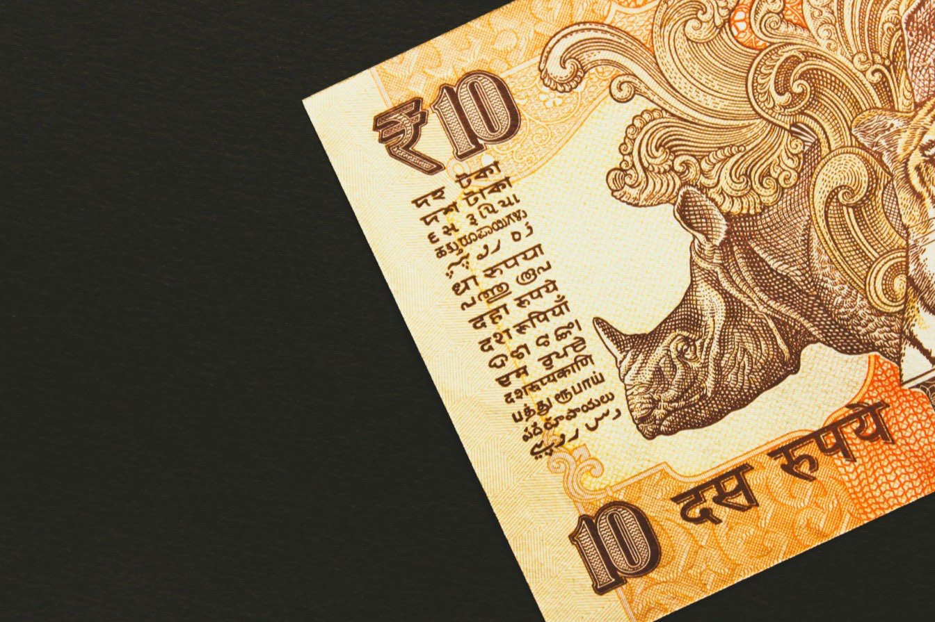 The corner of an Indian ten-rupee banknote on a dark surface, showing the rupee symbol, the denomination in the multi-language panel, and an engraved rhinoceros.